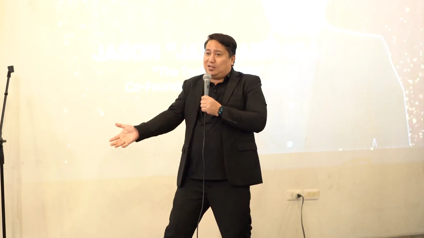 Jas Barinaga in a black suit, speaking with a handheld microphone.